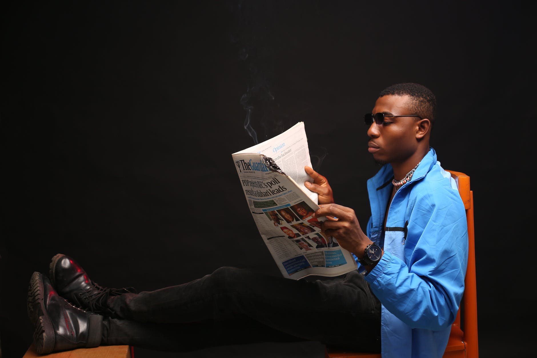 Photo of a young man with short hair and wearing a long sleeved light blue sports jacket, black pants and boots and sunglasses, sitting on a chair with his feet up on a stool, reading a newspaper.